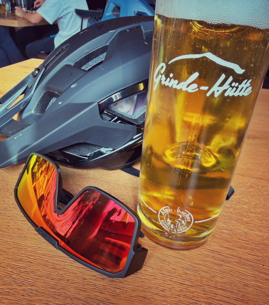 Ein Bierglas mit Grinde-Hütte-Aufdruck neben Helm und Sonnenbrille auf einem Holztisch