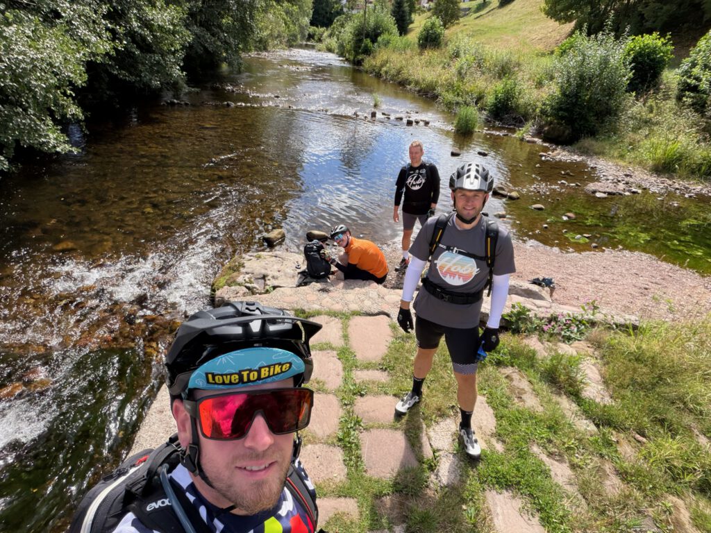 Vier Radfahrer an einem flachen Fluss, einer kniet am Ufer