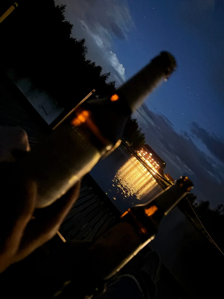 Zwei Bierflaschen stoßen bei Dämmerung an, im Hintergrund die beleuchtete Uferpromenade am See